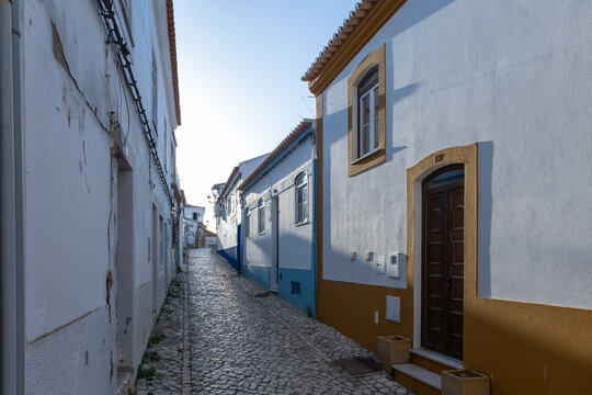 Narrow cobblestone street with whitewashed houses and blue and ochre trim in Ferragudo, Portugal