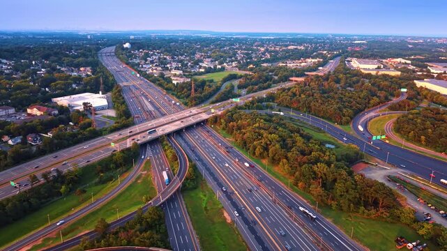 Approaching the huge multi-lane highway with multiple moving cars. There are green trees, grassy fields, houses, and some white commercial buildings on the sides of the roads.