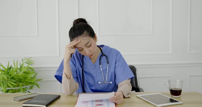 Exhausted female doctor in scrubs review read analyze paperwork at a desk, surrounded by medical equipment, notebooks, tablet, and coffee. burnout, and mental pressure in a clinical setting.
