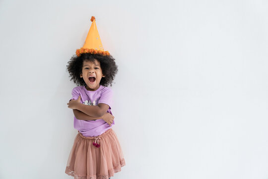 Portrait of happy young African black toddler little girl wearing a colorful party hat, purple shirt, pink skirt, and patterned tights, posing for a cheerful birthday halloween day celebration.