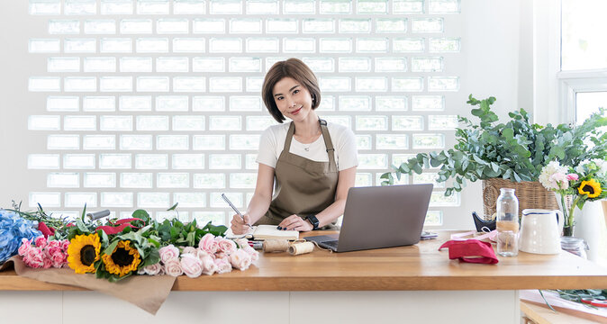 Startup small business, asian woman business owner home office, online marketing delivery,  e-commerce telemarketing. Florist celebrating success write notebook laptop flowers in  floral workspace