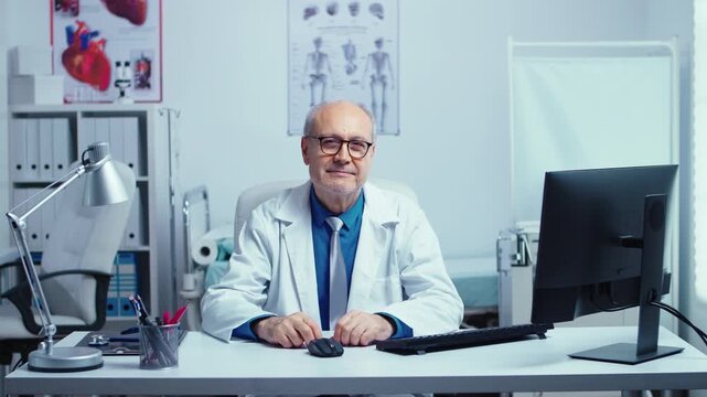 Senior male physician sits at a computer workstation and smiles toward camera in a bright medical office, presenting approachable professional healthcare, supporting digital workflow.