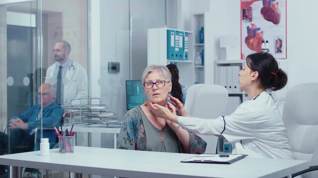 Older woman touches her neck while speaking with a female doctor in a modern clinic, supporting symptom discussion, primary care, medical assessment and patient communication during the visit.
