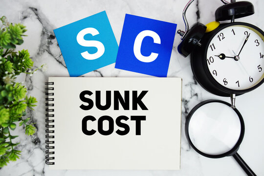 SC Sunk Cost text written on notepad with alarm clock and magnifying glass on marble background