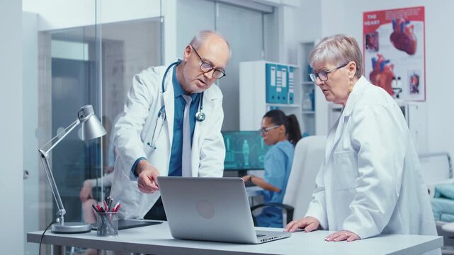 Two experienced doctors stand beside a laptop in a modern clinic, discussing clinical information and coordinating patient care, emphasizing clinical teamwork and collaborative care.