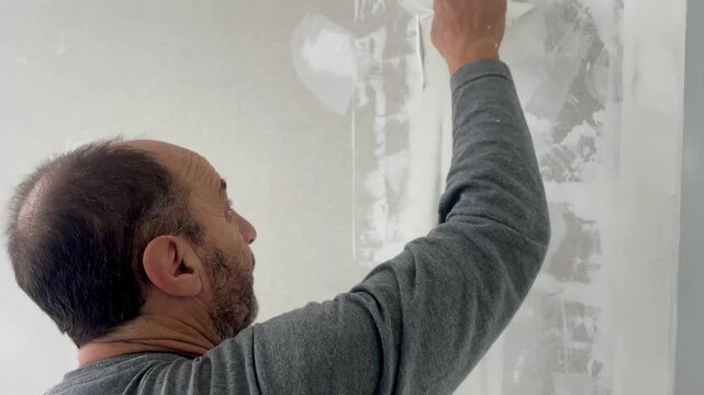 Senior man spreading white joint compound over drywall with a putty knife to prepare the wall for finishing.