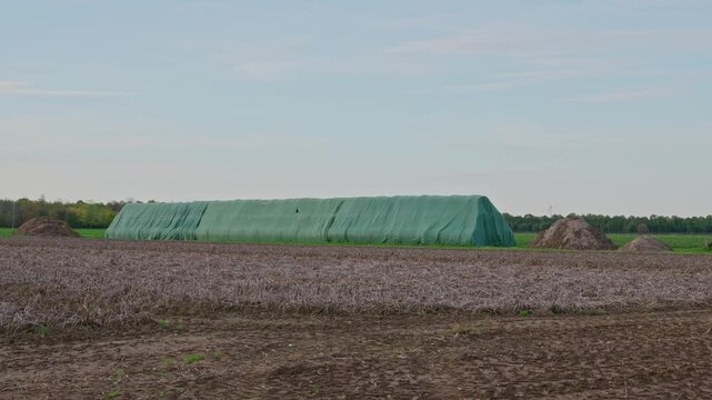 Wide panoramic view of an agricultural field with a large silage storage covered with a green plastic tarp in a rural landscape under a cloudy sky