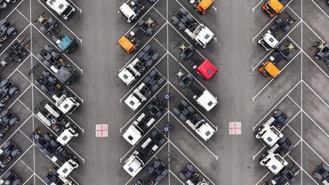 Aerial view of rows of parked new trucks and commercial vehicles in a large paved parking lot with colorful cabs and marked spaces. Drone footage of warehouse of manufactured lorry cars. Industrial co