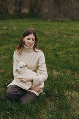 Naklejka premium Bright outdoor scene with a woman in a cozy cream sweater sitting in a grassy field, gently holding a white goat kid, capturing a peaceful, tender lifestyle moment in sunlight