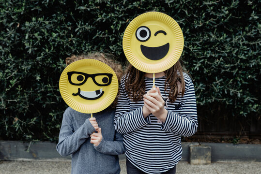Anonymous brother and sister standing together holding handmade happy yellow emoji / emoticon masks; Melbourne, Victoria, Australia
