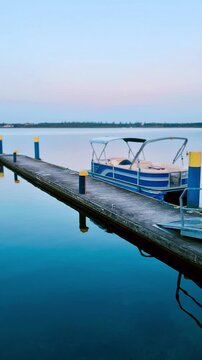 A motorized pontoon boat with a canopy is moored to a concrete dock at dusk. 