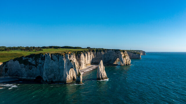 White limestone cliffs rise above the blue sea at Etretat, with a natural arch and a sea stack near the shoreline under a clear sky.