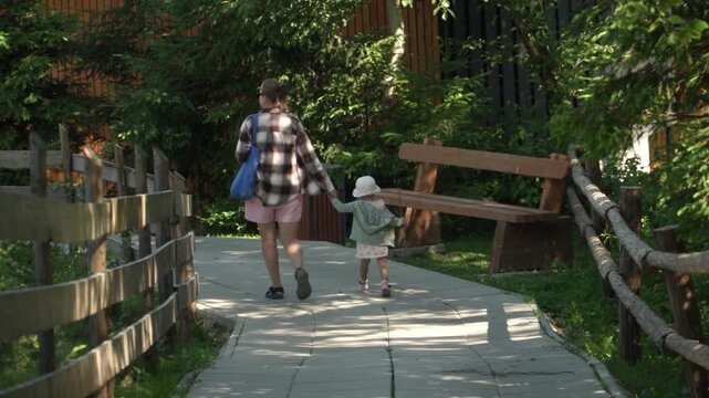 A little girl pink dress carries bundle sticks, holding mother hand look concentration, shot behind showing family strolling along paved path leading down through woods toward wooden houses warm