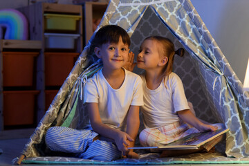 Childhood imagination and friendship, little girl whispering a secret to her brother inside a cozy play tent at home during bedtime © Pixel-Shot