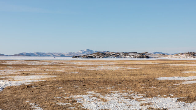 Panoramic view of a vast winter steppe landscape with dry yellow grass and snow patches leading to frozen Lake Baikal in Irkutsk Oblast, Russia under a clear blue sky on a cold sunny day