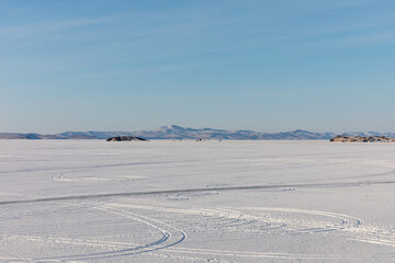 Wide scenic view of the snow-covered frozen expanse of Lake Baikal with tire tracks and distant mountain ridges in Irkutsk Oblast, Russia under a clear sky © TravelFlow