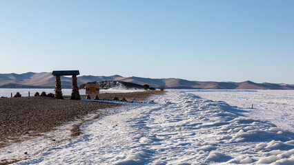 Scenic view of the stone portal on Cape Uyuga spit beside frozen Lake Baikal in Irkutsk Oblast, Russia during cold winter with snowy shores and distant barren hills under clear blue sky © TravelFlow