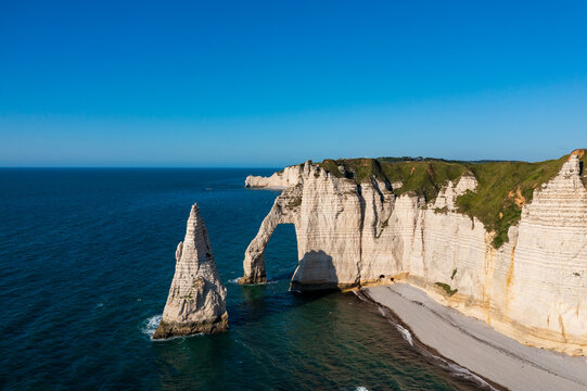 White chalk cliffs, a natural sea arch and a rock stack overlook the blue English Channel at Etretat. A pebble beach and coastal headland lie under a clear sky.