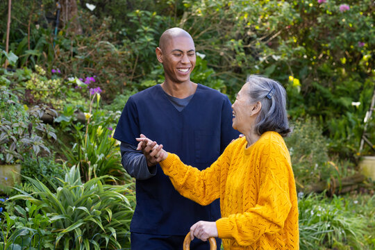 Caregiver in navy-scrubs guiding senior Asian woman in mustard-sweater using cane on garden path