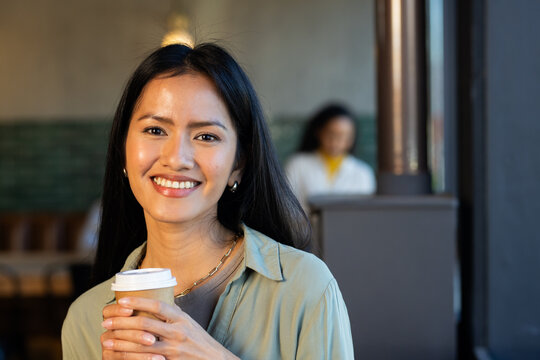 Mid-adult Asian woman smiling, holding takeaway cup with white lid at cafe window, wearing necklace