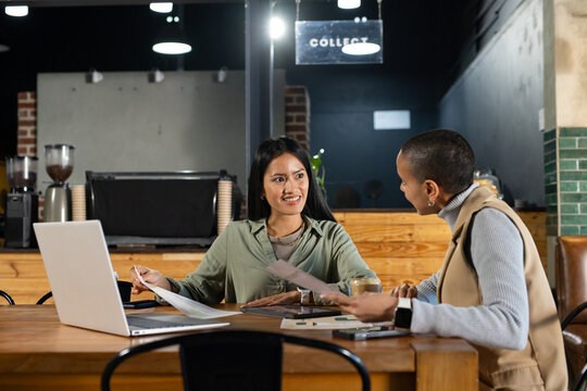 Asian woman and African American woman reviewing documents at cafe with laptop, pens, coffee