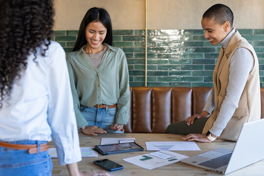 Diverse female colleagues in casual workwear standing and reviewing charts, tablet at meeting table
