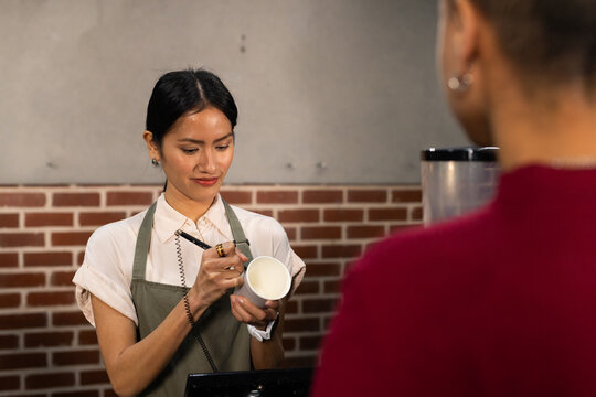 Asian barista in green apron labeling cup with marker serving African American customer at cafe POS