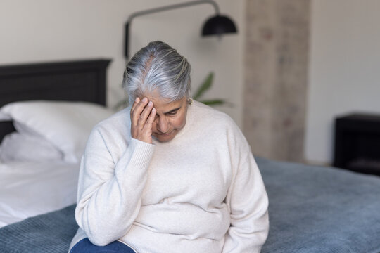 Senior African woman sitting on bed in bedroom wearing cream sweater, pressing forehead