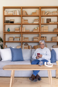 Senior woman sitting on gray sofa holding red smartphone near coffee table, bookshelf, copy space