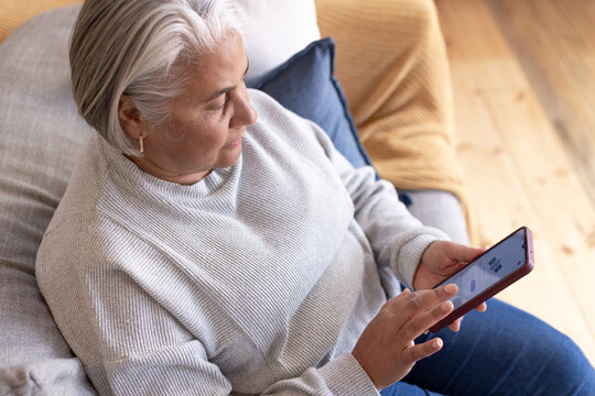 Senior African-American woman sitting in gray sweater on couch at home holding purple smartphone