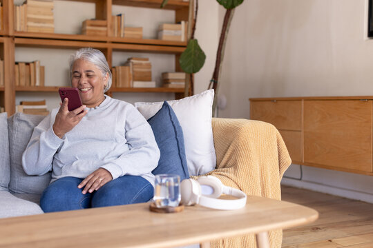 Mature African American woman sitting on gray sofa at home wearing sweater, holding maroon phone