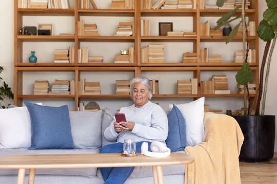 Senior woman of African heritage sitting on gray sofa holding red smartphone by wooden bookcase