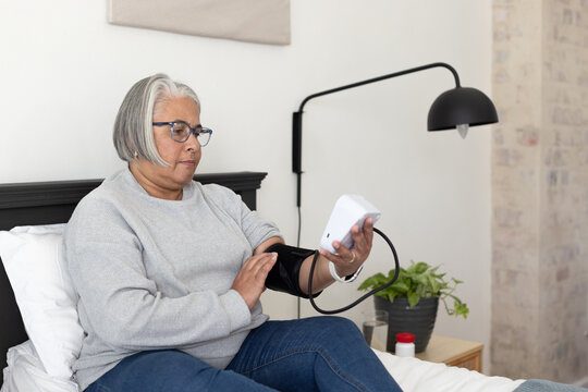 Senior African American woman sitting on bed in bedroom checking digital monitor with black cuff