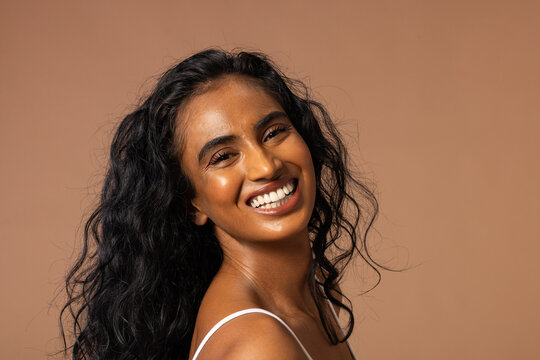 Woman smiling broadly while posing in studio on beige backdrop wearing white spaghetti-strap top
