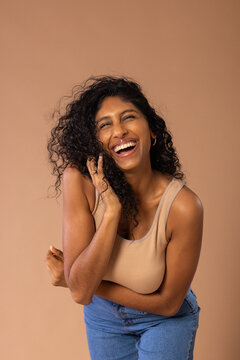 Indian woman laughing, touching hair in studio wearing beige ribbed top blue jeans, gold hoops