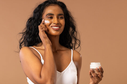 Female model wearing white camisole applying cream to right cheek at studio, holding jar, smiling