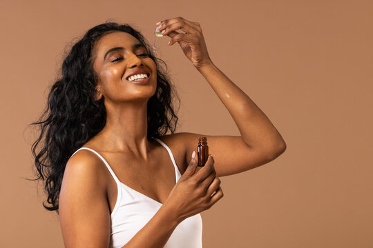 Indian woman wearing white tank top applying serum with amber dropper above forehead in studio