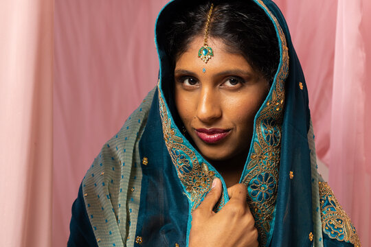 Indian woman touching teal embroidered dupatta at pink backdrop, posing with maang tikka bindi