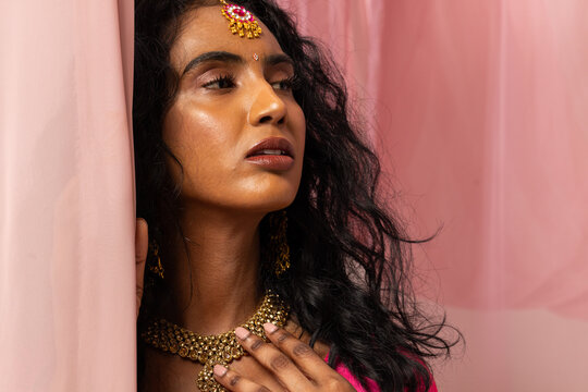 Adult Indian woman posing in studio wearing bright pink garment and maang tikka, touching curtain