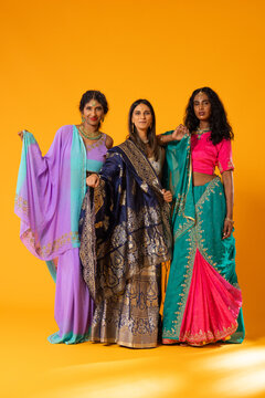 Diverse female friends posing against orange backdrop wearing embroidered saris and gold jewelry