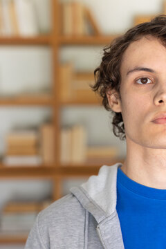 Man facing camera sitting in study with wooden bookshelf wearing blue shirt gray hoodie, copy space