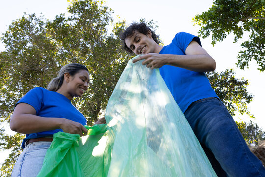 Male and female volunteers holding large green trash bag in leafy park wearing matching blue tees