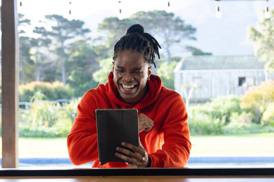 Adult African man in orange hoodie laughing, holding tablet in folio at counter under string lights