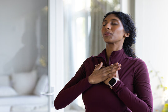 African American woman standing eyes closed, breathing at home near window, smartwatch, copy space