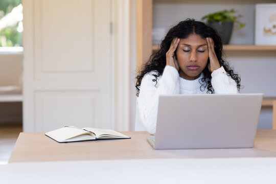 African American woman in white knit holding temples at home desk with open laptop, notebook