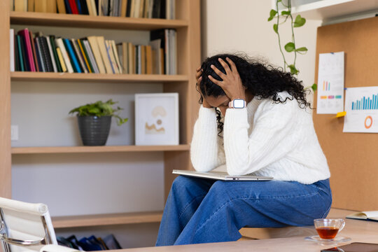 African American woman sitting on desk in study holding head with hands over laptop, copy space