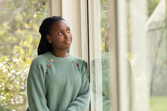African woman standing by glass window with reflection wearing sage sweater gazing up, copy space