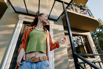 Fototapeta premium Young woman wearing a green knit sweater and orange jacket stands outside a modern home, smiling confidently. Casual fashion concept for lifestyle and streetwear.