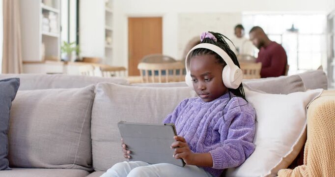 African American child sitting on gray sofa wearing headphones, tapping tablet at clip, smiling