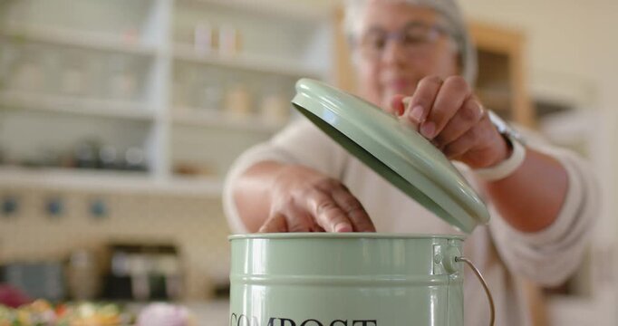 Woman lifting green compost lid, dropping red onion peel into pail for meal prep at counter